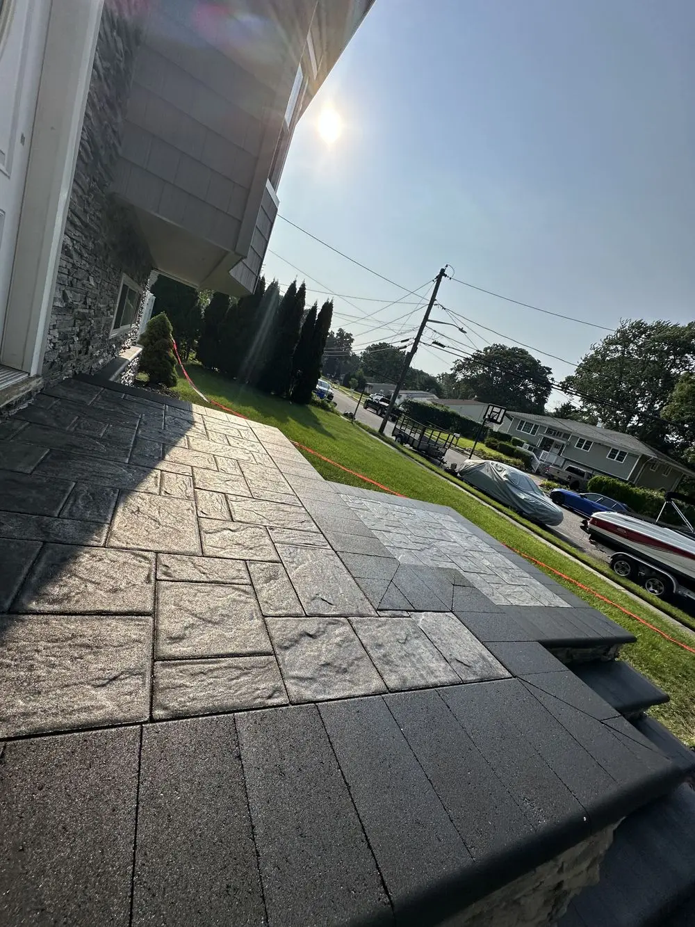 A sunlit, textured stone porch leads to a residential street. There are neatly trimmed bushes and a lawn in the yard. Houses and parked cars line the street under a clear blue sky.