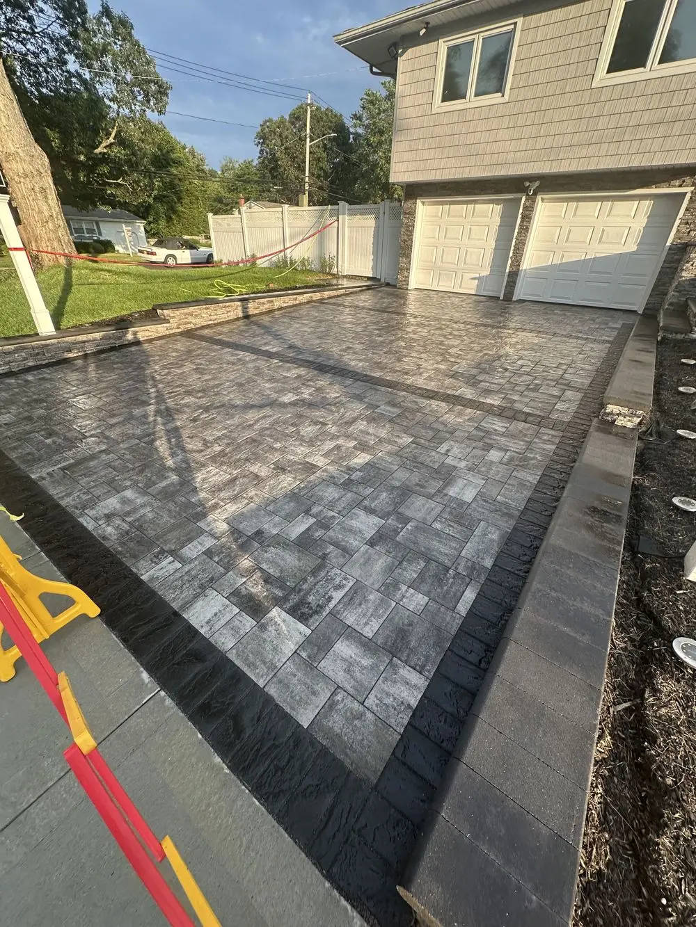A residential driveway with interlocking gray and black brick paving. The driveway is bordered by black bricks and leads to a garage with two white doors. A portion of the yard and a white fence are visible in the background.