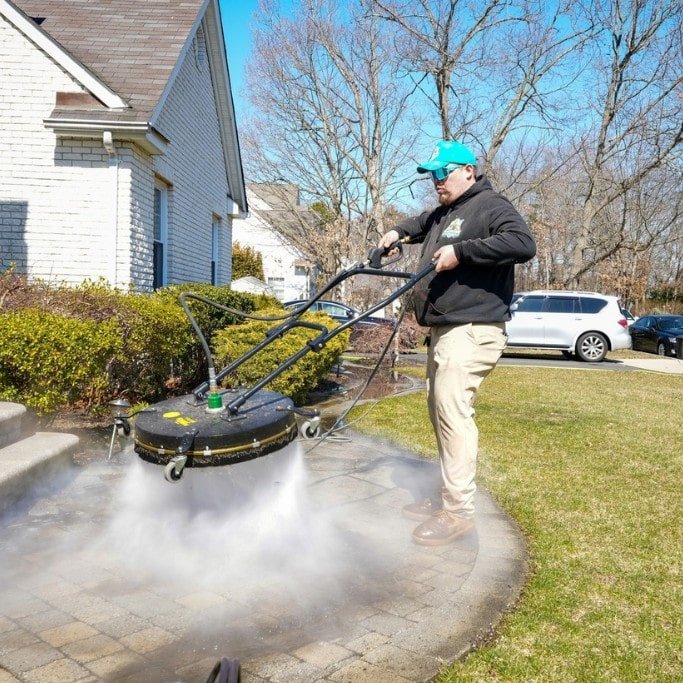 A person wearing a turquoise cap and black hoodie is using a power washer to clean a paved pathway in front of a white brick house. The yard has green grass, bare trees, and a few bushes. A white car is parked in the driveway.