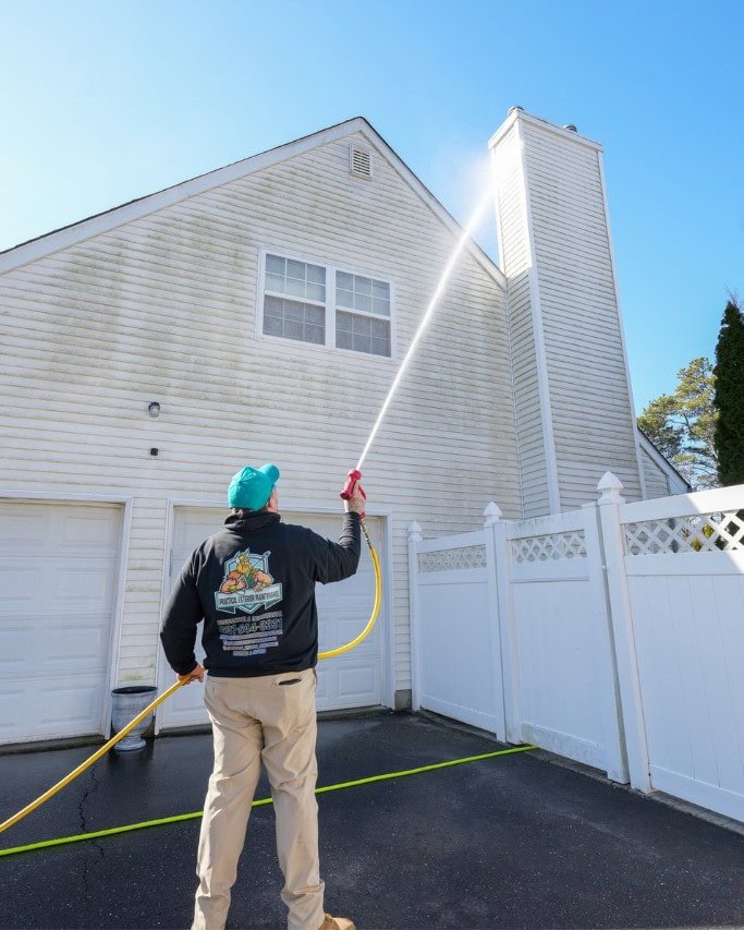 A person in a black jacket and beige pants uses a hose to power wash the exterior of a white house with a tall chimney. A section of a white fence and a closed garage door are visible. The sky is clear and blue.