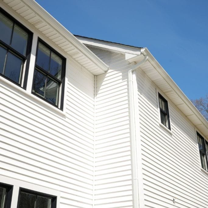 Close-up view of a house exterior featuring white siding and black-framed windows against a clear blue sky. The image captures the side and roofline of the building.
