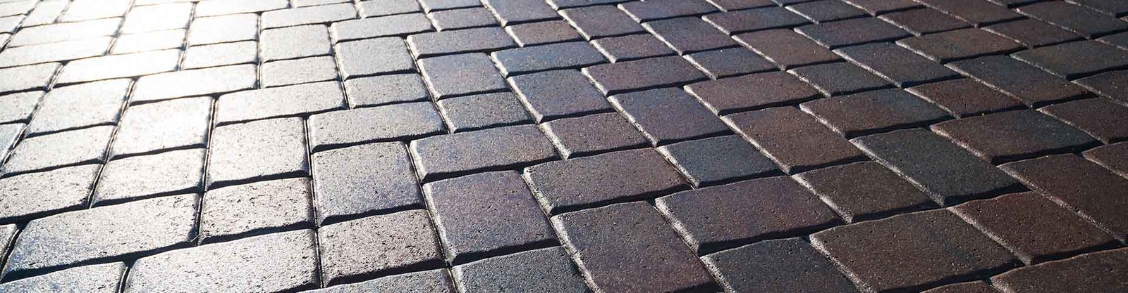 Close-up of a surface made up of interlocking stone pavers. The stones are square-shaped and in varying shades of gray and brown, creating a uniform pattern. The lighting gives a slight sheen to the stones, accentuating their texture.