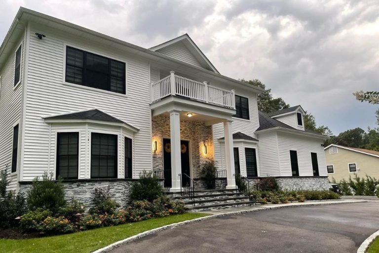 A large two-story white house with black-framed windows, a stone facade, and a columned entrance. It features a balcony, a curved driveway, and landscaped bushes and flowers in front. Overcast sky in the background.