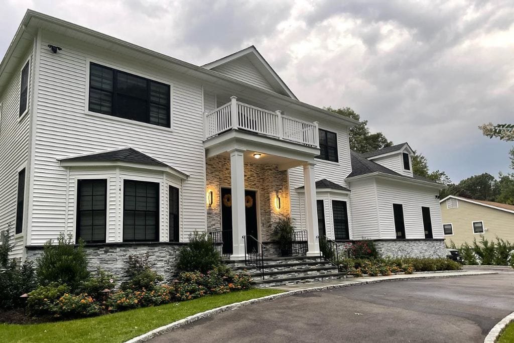 A large two-story white house with black-framed windows, a stone facade, and a columned entrance. It features a balcony, a curved driveway, and landscaped bushes and flowers in front. Overcast sky in the background.