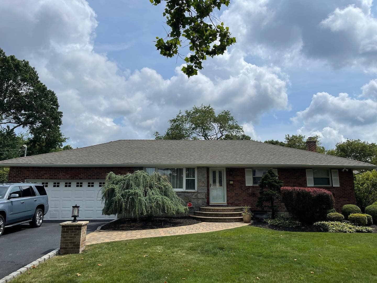 A single-story brick house with a gray roof features a small porch and circular steps leading to the front door. A neatly manicured lawn and garden with shrubs and trees surround the house. A black SUV is parked in the driveway. Clouds fill the sky.