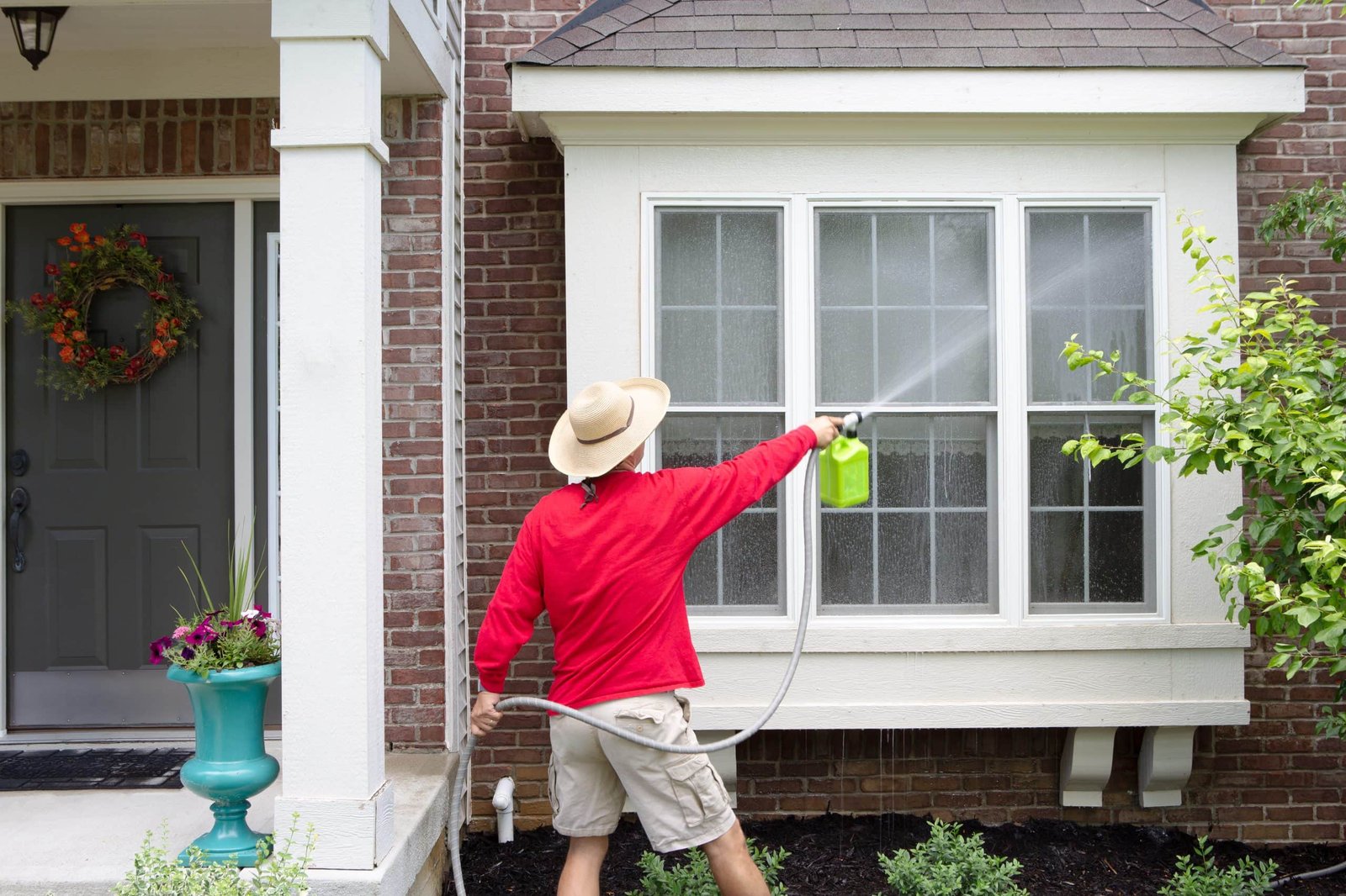 A person in a red shirt and straw hat is power washing a large window on a brick house. They are holding a green spray nozzle attached to a hose. A planter with flowers stands beside a door adorned with a floral wreath.