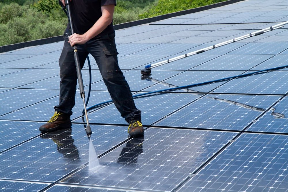 A person is cleaning solar panels on a roof using a high-pressure water hose. They are wearing dark clothing and yellow-laced boots. A long cleaning pole is visible on the panels, and trees are in the background.