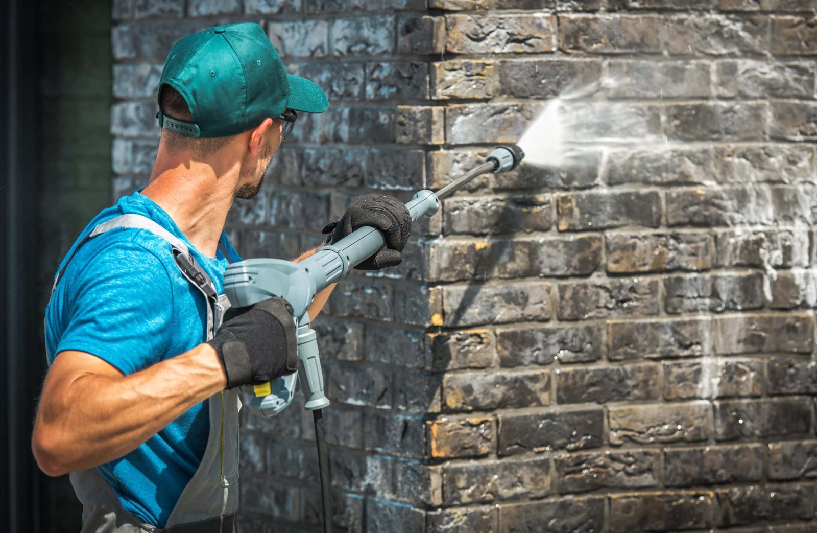 A man in a blue shirt and cap, wearing gloves, uses a pressure washer to clean a brick wall. The wall is covered with grayish residue as water sprays from the washer, highlighting the cleaning process.