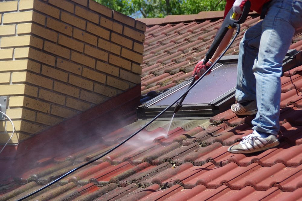 Person power washes a red tile roof near a brick chimney and a skylight, removing dirt and debris. They are wearing blue jeans, a red shirt, and white sneakers. The sky is clear and sunny.