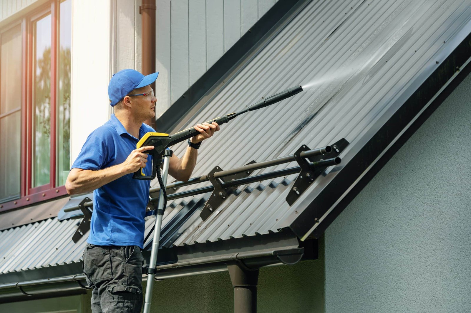 Person in a blue shirt and cap using a power washer to clean a metal roof. They are standing on a ladder, directing the water spray onto the roof panels.
