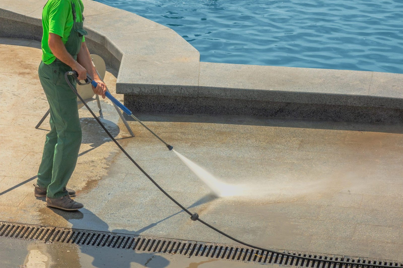 A person in green shirt and overalls uses a pressure washer to clean a stone surface near a pool. Water sprays from the nozzle, and the ground appears wet. The pool's blue water is visible in the background.