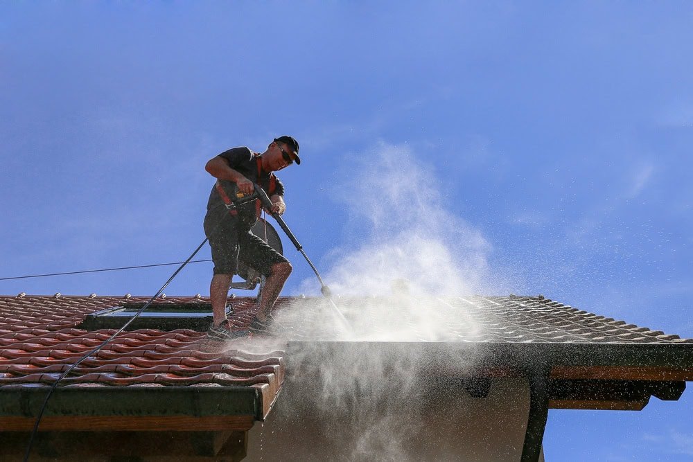 A person wearing a cap stands on a tiled roof, power washing it. Water sprays over the tiles, creating a mist against the clear blue sky. Safety ropes are attached to the roof, ensuring security during the cleaning process.