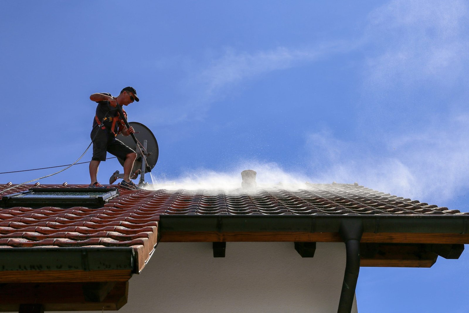 A person in safety gear stands on a tiled roof, using a pressure washer to clean it. Water sprays into the air, creating a mist under a clear blue sky.