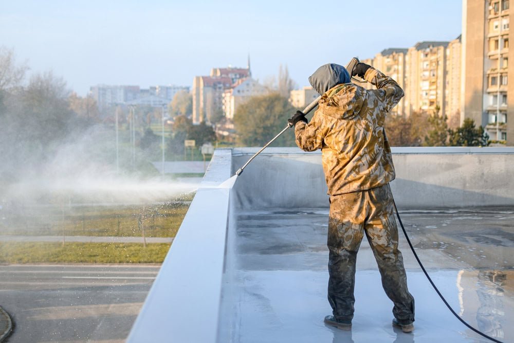 Person in camouflage clothing pressure washing a flat rooftop. The individual wears a hood and stands near the edge, spraying water over the roof surface. Urban buildings and a blurred background of trees and a road are visible beyond the roof.