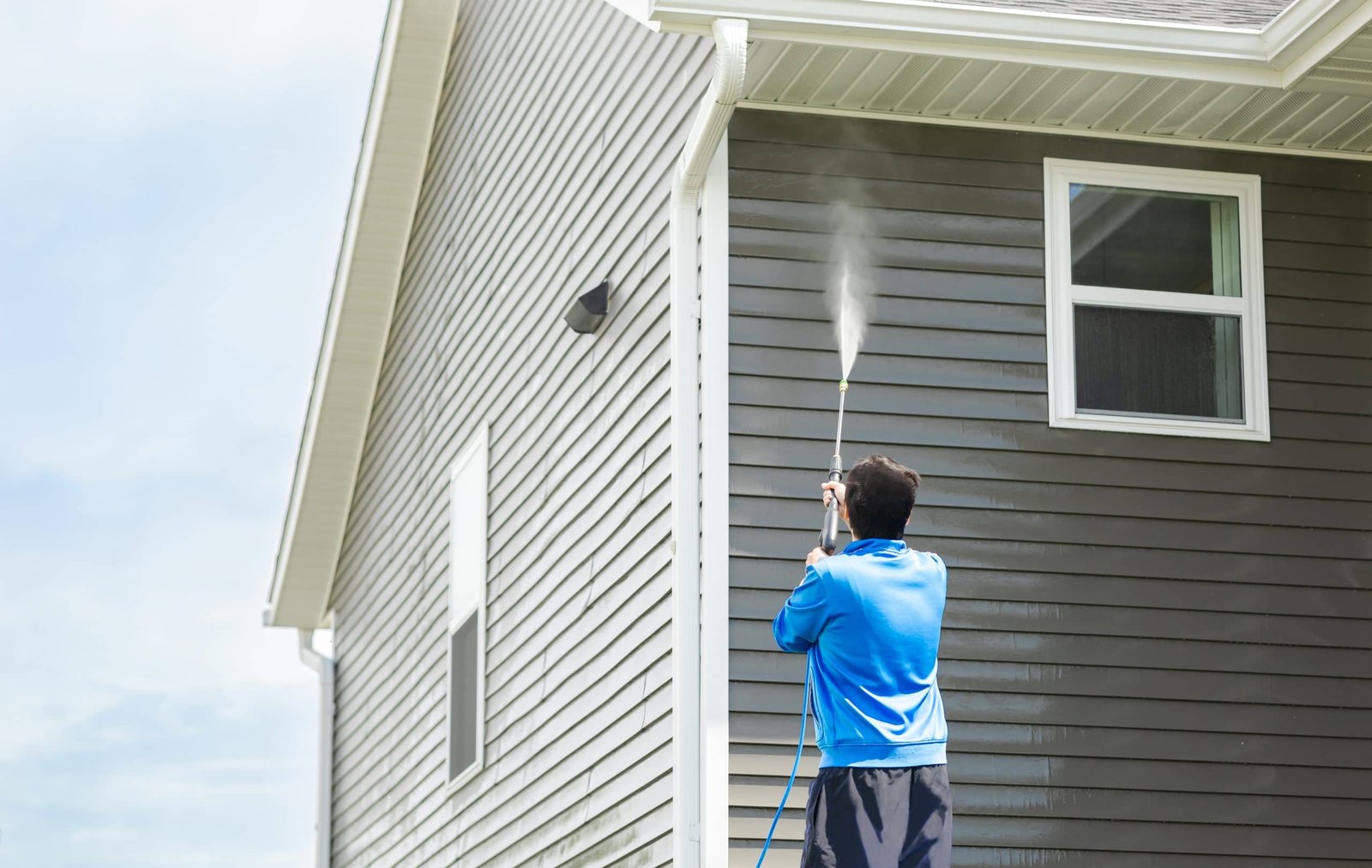 A person in a blue hoodie power washes the exterior of a house with gray siding. The person is facing away, and water is seen spraying onto the upper part of the house. There is a single window visible on the wall.