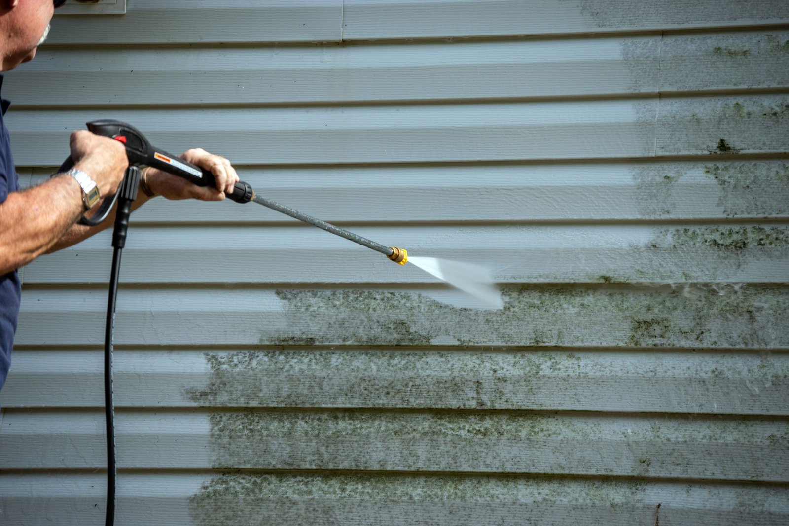 A person using a power washer to clean the exterior siding of a building. The left side of the wall appears clean, while the right side is still covered with dirt and grime. The person is holding the washer with both hands.