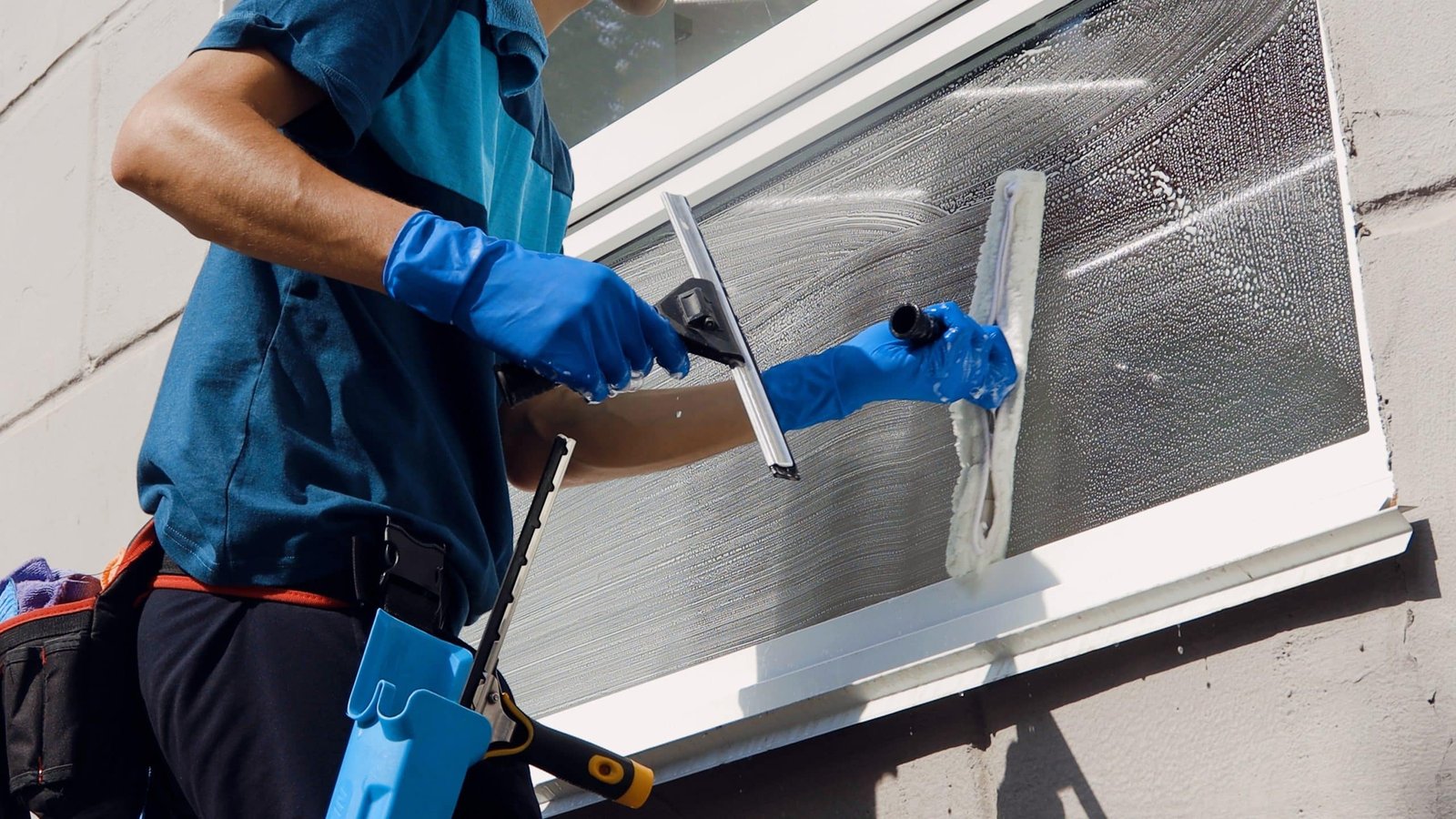 A person wearing blue gloves is cleaning a window with a squeegee and a scrubber. They are dressed in a blue uniform and have cleaning tools attached to their belt. The window is partially covered with cleaning solution.