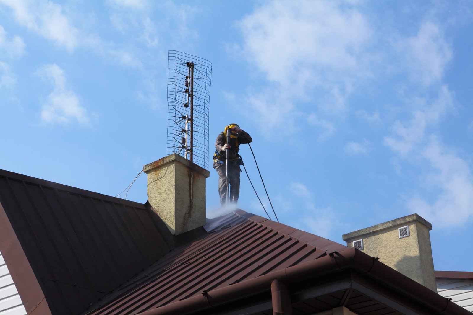 A worker wearing protective gear power washes the metal roof of a house on a bright day. They are secured with safety ropes. An antenna is visible on a chimney close by. The sky is clear with a few clouds.