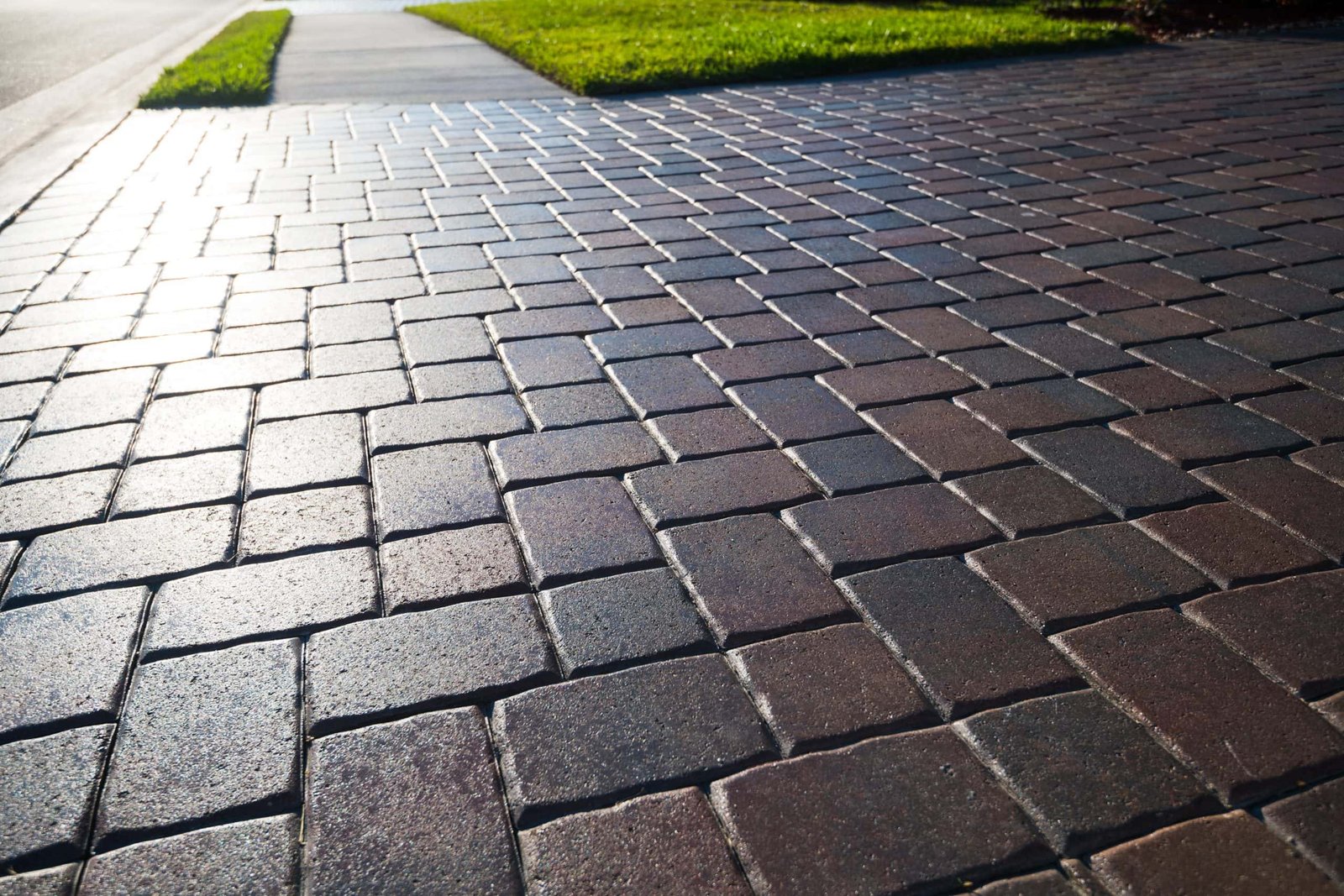 Brick pavement with a sunlit glow, leading to a patch of green grass on the right. The bricks are arranged in a staggered pattern, and the sidewalk extends into the distance under clear skies.