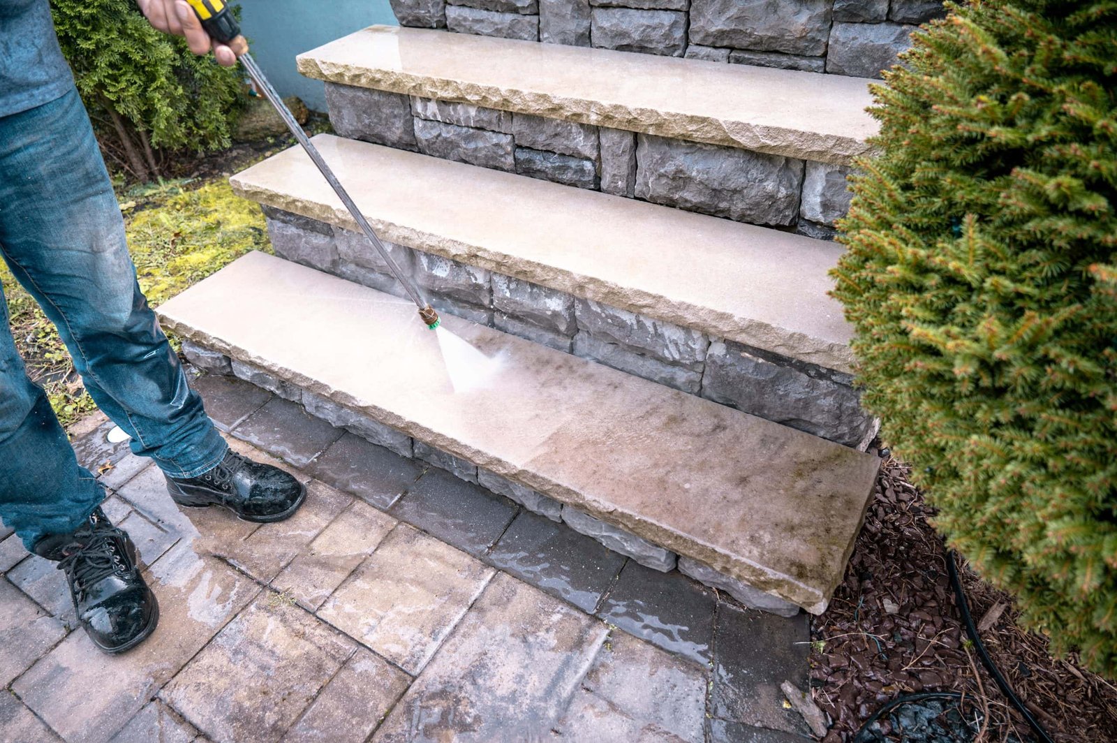 A person is power washing stone stairs. The jet of water is cleaning the grime from the surface. The steps lead up from a paved patio, and there is greenery, including a bush, visible on the side. The person is wearing jeans and black shoes.