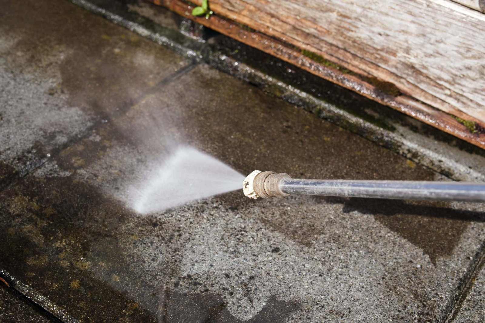 A high-pressure water jet cleaning a concrete patio, removing dirt and grime. The spray creates a mist as it hits the surface, and the edges of the patio are bordered by a wooden structure.