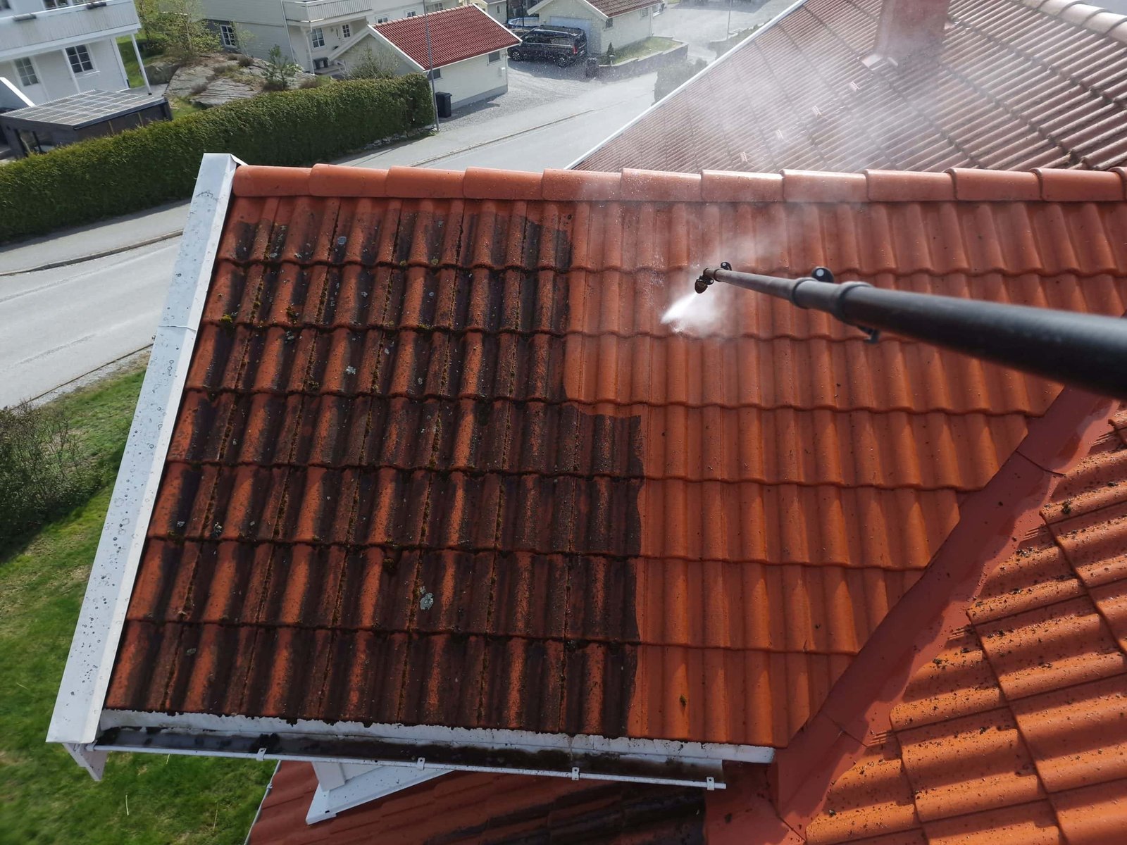 A person is using a pressure washer to clean a red tiled roof. The left side of the roof is dirty with black stains, while the right side is visibly cleaner. Residential houses and a road are in the background.