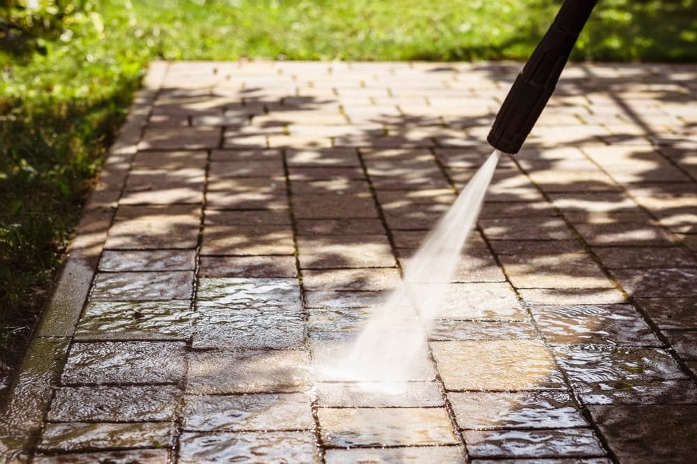 A pressure washer is cleaning a stone patio. Water is being sprayed onto the surface, removing dirt and grime. The patio is arranged in a grid pattern, with sunlight and shadows visible on the wet and dry areas.