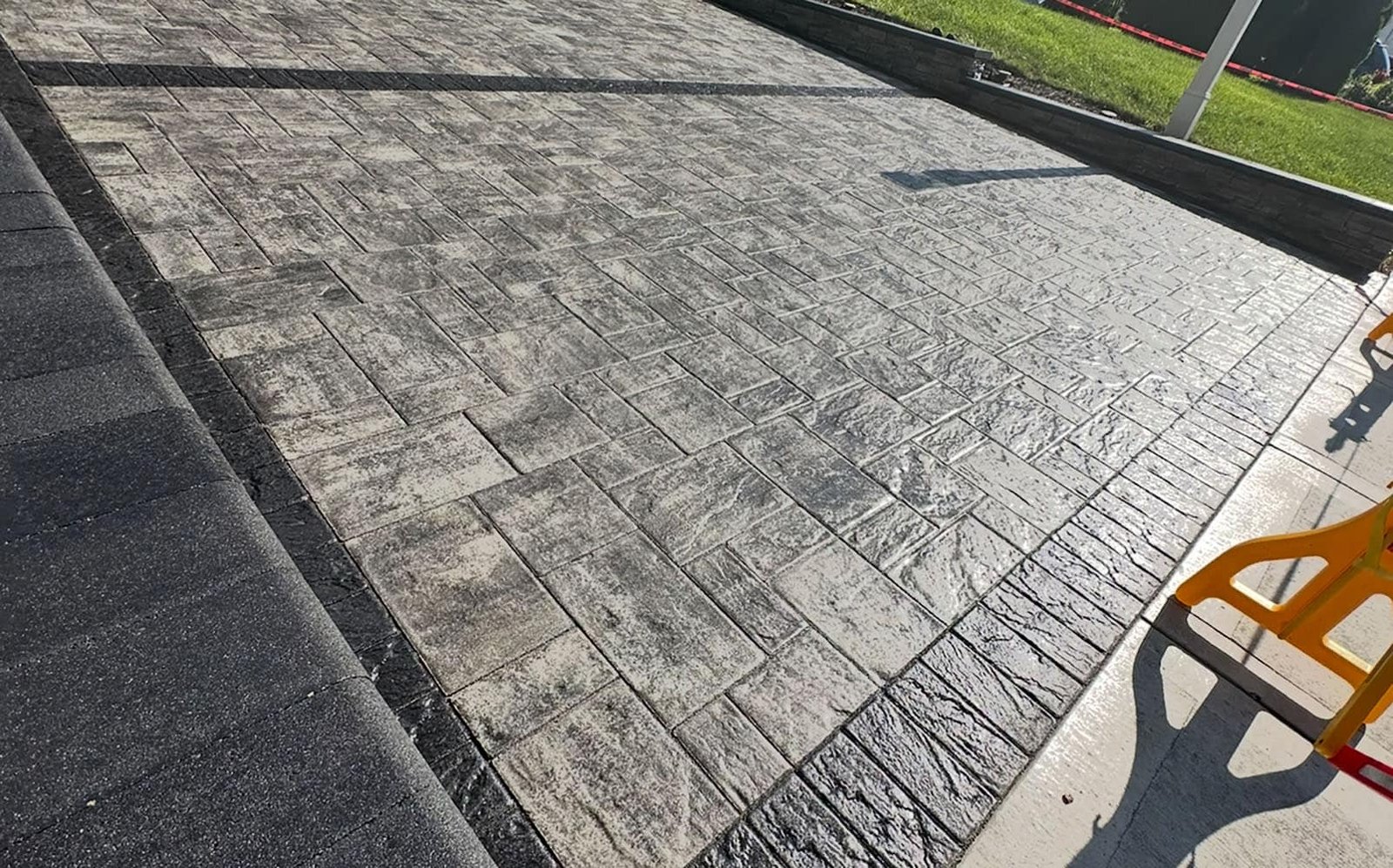 Freshly laid patterned concrete paving with a brick-like design is shown, glistening under sunlight. A shadow of a light pole stretches across the surface, which is bordered by grass on one side and concrete on the other.