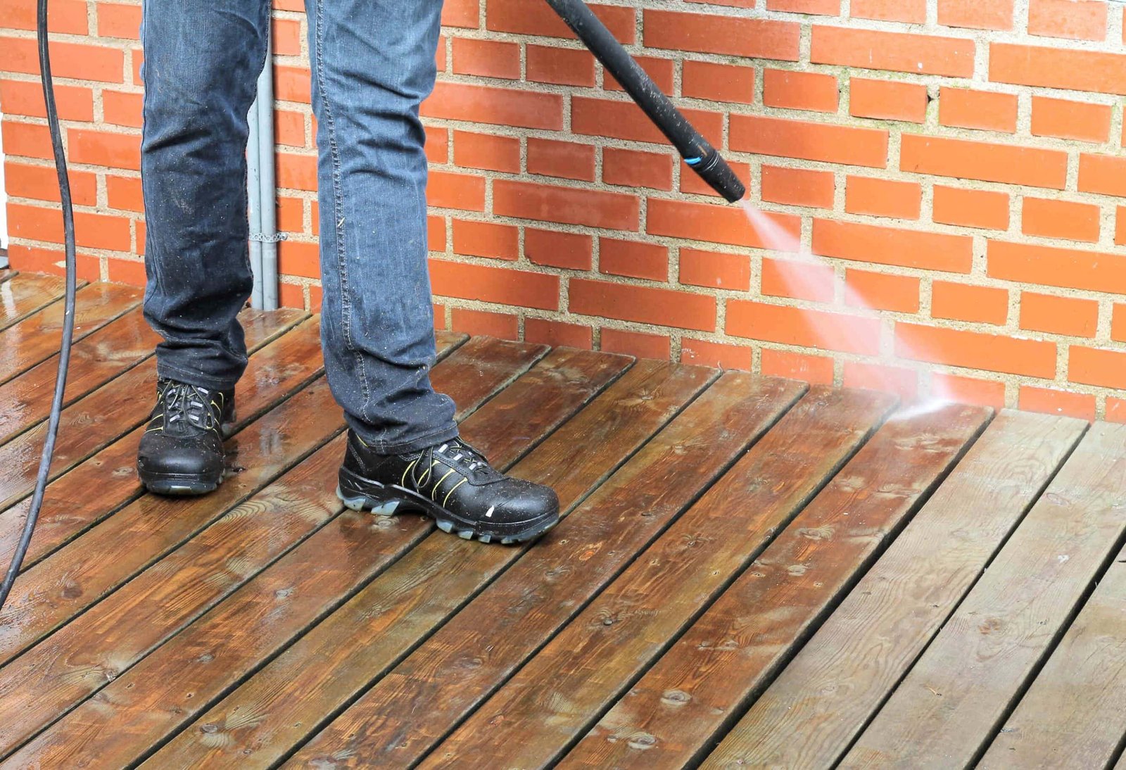 A person using a pressure washer to clean a wooden deck. They are wearing jeans and black boots. The water is visibly spraying onto the deck, removing dirt, with a red brick wall in the background.