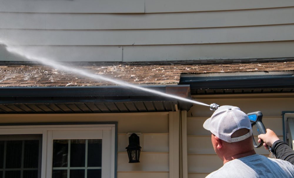 A person in a white cap is pressure washing the gutters of a house. They are using a spray nozzle to clean the area, with water spraying upward onto the roof edge. The house exterior is beige with a black outdoor light fixture visible.
