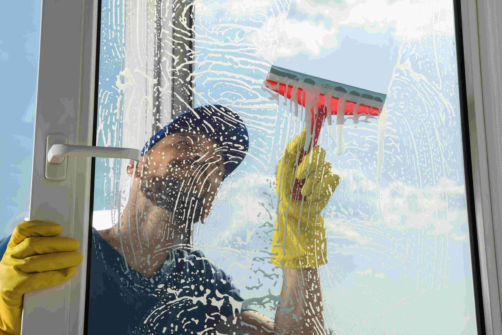 A person wearing yellow gloves and a blue cap cleans a large window with a red squeegee. The window is covered in soap suds, revealing a clear blue sky with clouds behind it.