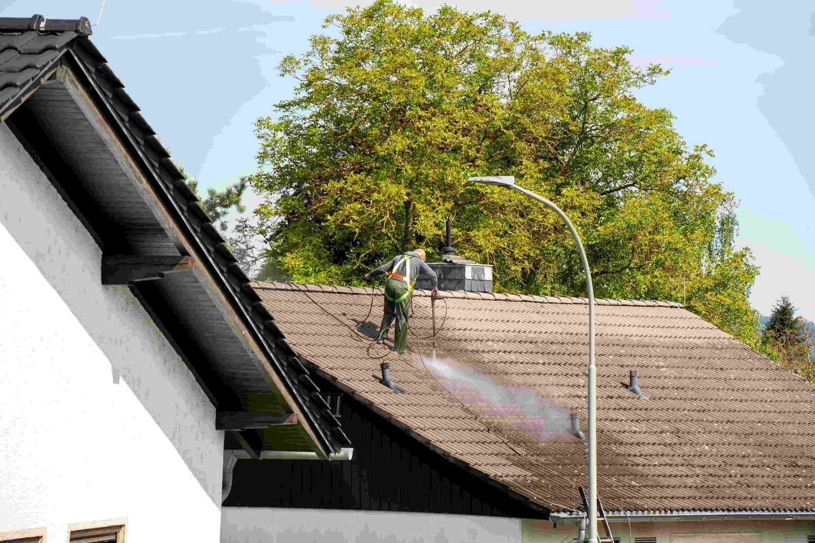 A person stands on a house roof using a hose to spray water or cleaner on the shingles. There is a tree and a streetlamp nearby. The sky is clear and the roof is partially wet.