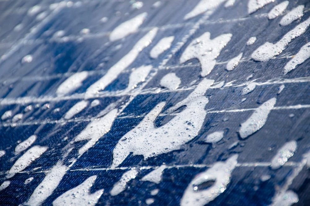 Close-up of a solar panel with water and soap suds covering its surface. The blue of the panel contrasts with the white soap bubbles, creating an abstract pattern. The image suggests cleaning and maintenance of solar equipment.
