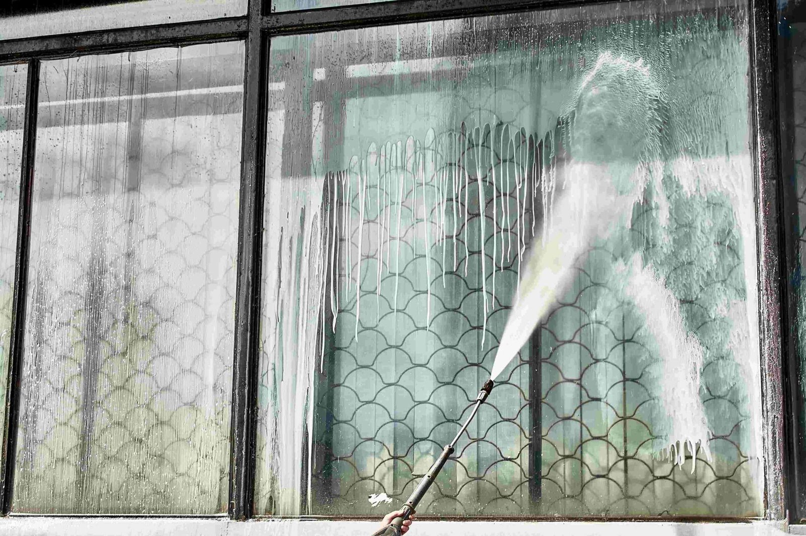 A person uses a high-pressure hose to clean a large, gridded glass window, removing grime and revealing a clear pattern beneath. Water splashes and drips down the window as it's being washed.