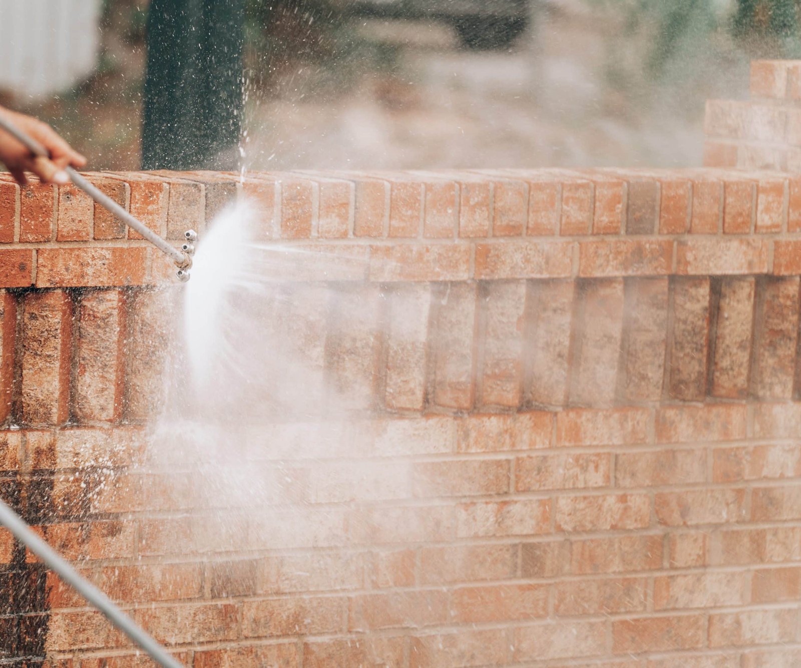 A person is using a pressure washer to clean a brick wall. Water sprays forcefully against the bricks, creating mist and droplets in the air. The movement is dynamic, indicating the cleaning action.