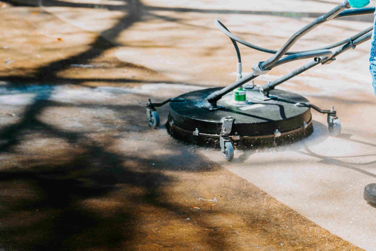 A person uses a circular pressure washing surface cleaner to clean an outdoor concrete area. The equipment sprays water, removing dirt and grime. Shadows from nearby trees are visible on the ground.