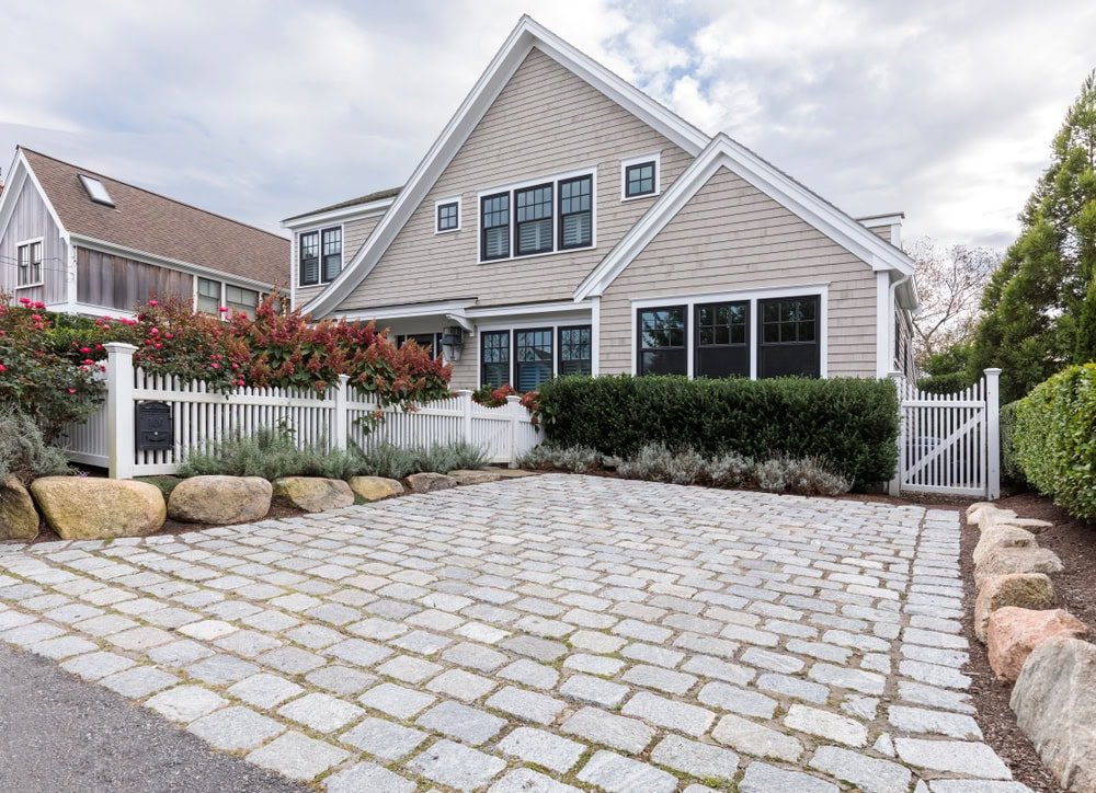 A modern beige house with black-trimmed windows, white picket fence, and a cobblestone driveway surrounded by lush landscaping and flowering bushes under a cloudy sky.