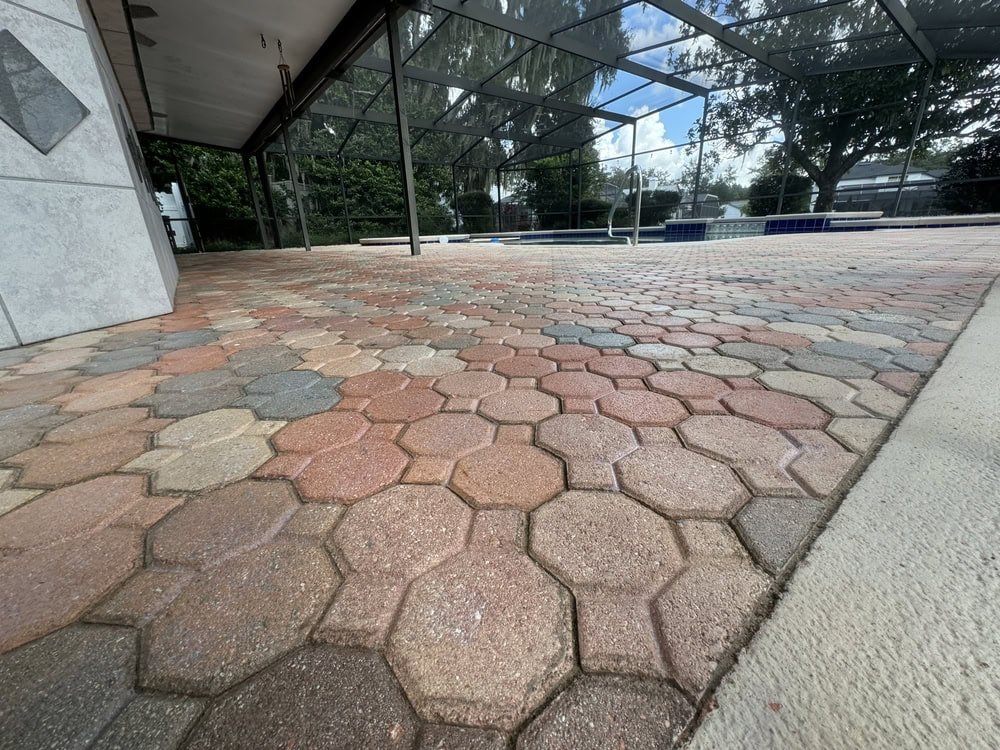 Close-up view of a multicolored hexagonal paver patio next to a swimming pool, with a screened enclosure and trees visible in the background under a partly cloudy sky.