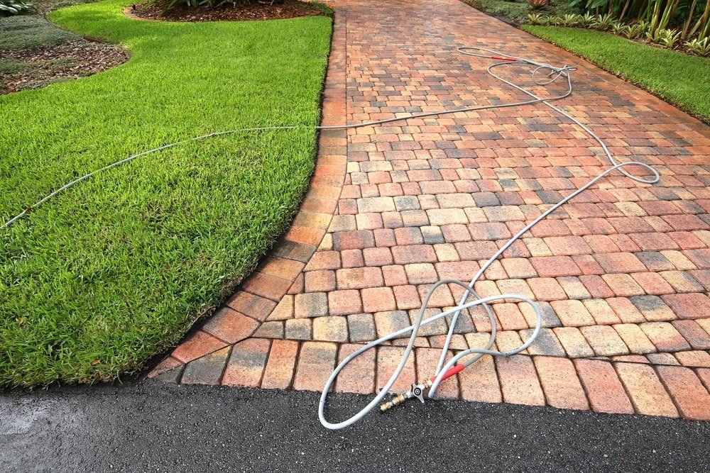 A long garden hose is stretched across a brick driveway and grassy lawn, looping in several sections near the driveway’s edge. The hose appears to be attached to a spray nozzle.